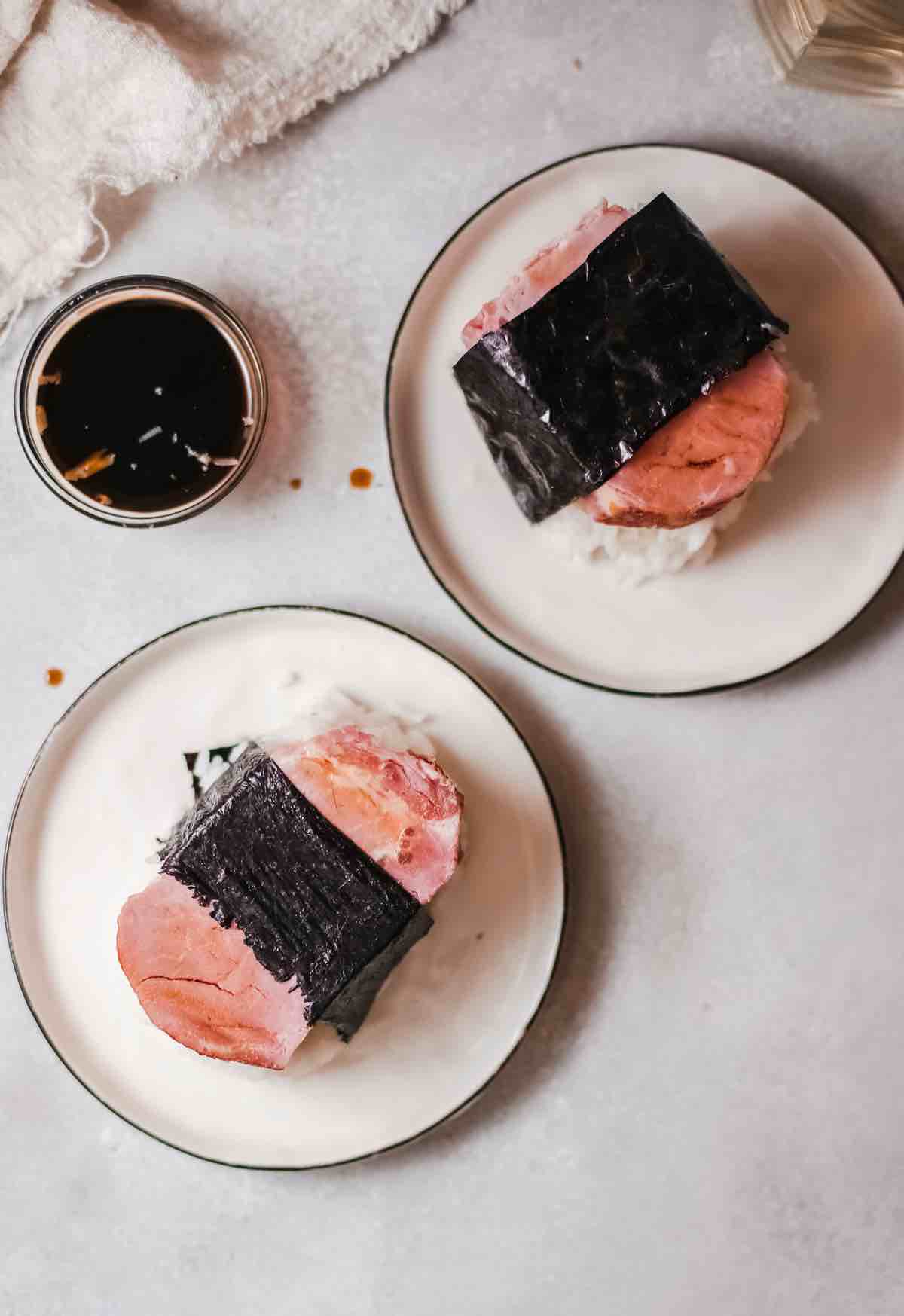 Learn how to make Spam Musubi by Rice Cooker Guy. Image of two Spam Musubis with a cup of sauce.